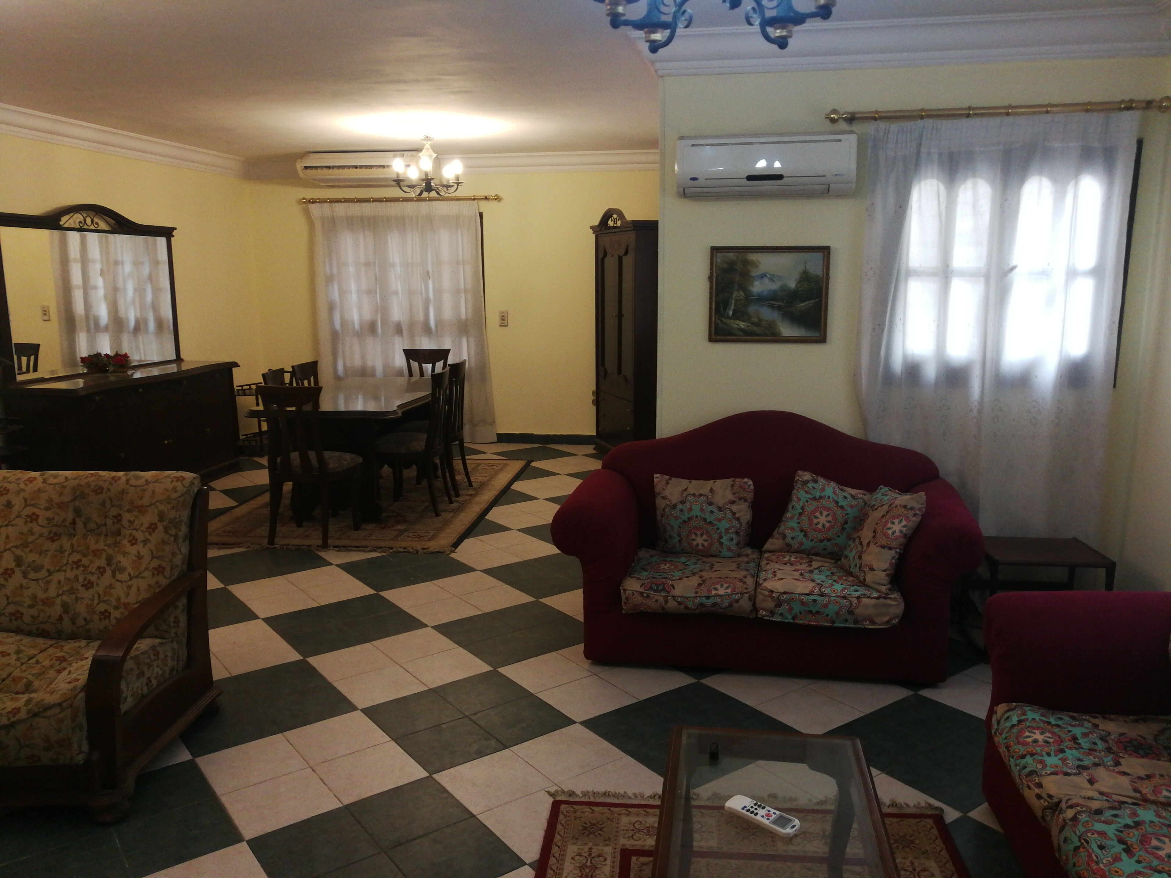 Ein Wohnzimmer mit Schachbrettmuster-Boden, einem roten Sofa, einem blumigen Sessel, einem Glastisch und einer Essecke mit Tisch und Stühlen.