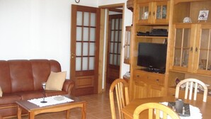 A living room with a brown leather sofa, a wooden coffee table, a dining table with chairs, and a television on a wooden cabinet.