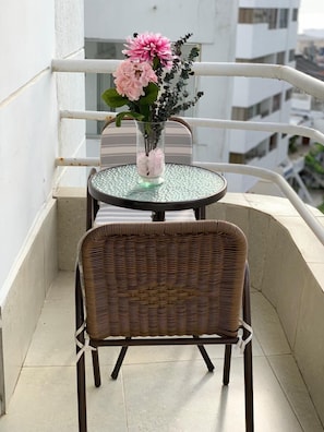 Un balcon avec une chaise en osier et une petite table ronde sur laquelle est posé un vase en verre contenant des fleurs roses.