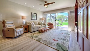 Un salon spacieux avec un canapé beige, un fauteuil inclinable et une table basse. On y trouve un ventilateur de plafond, une grande fenêtre donnant sur l’extérieur et un tapis à motifs sur le sol.