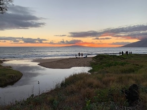 Cena de praia com um grupo de pessoas perto da água, um mar calmo e um pôr do sol ao fundo.