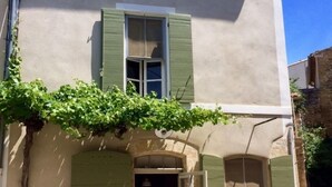 Un edificio a due piani con persiane verdi, una piccola area esterna con tavolo e sedie, e una porta che dà all'ingresso.