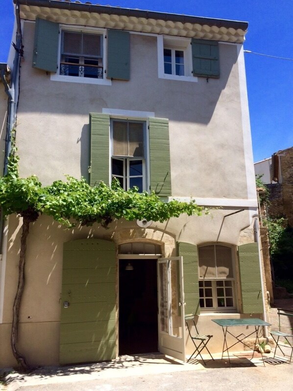 A two-story building with green shutters, a small outdoor seating area with a table and chairs, and a door leading to the entrance.