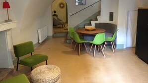 Un accogliente soggiorno con un tavolo rotondo in legno, sedie verdi, un poggiapiedi intrecciato e una scala che conduce a un'altra stanza.