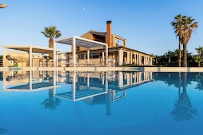 A pool with a clear reflection, a covered outdoor area with chairs, and a building with columns.