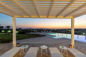 Un espacio exterior techado con piscina, sillones de descanso y vistas a la ciudad al atardecer.