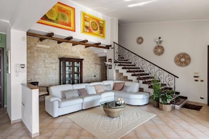 A modern living room with a sectional sofa, a glass coffee table, a staircase with a wrought iron railing, and a brick wall.