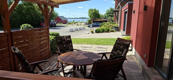 A patio with a round table and four chairs, a wooden fence, and a view of a road with cars and a building in the background. - Glewitz Am Alten Fährhaus 4 (Garz/Rügen)
