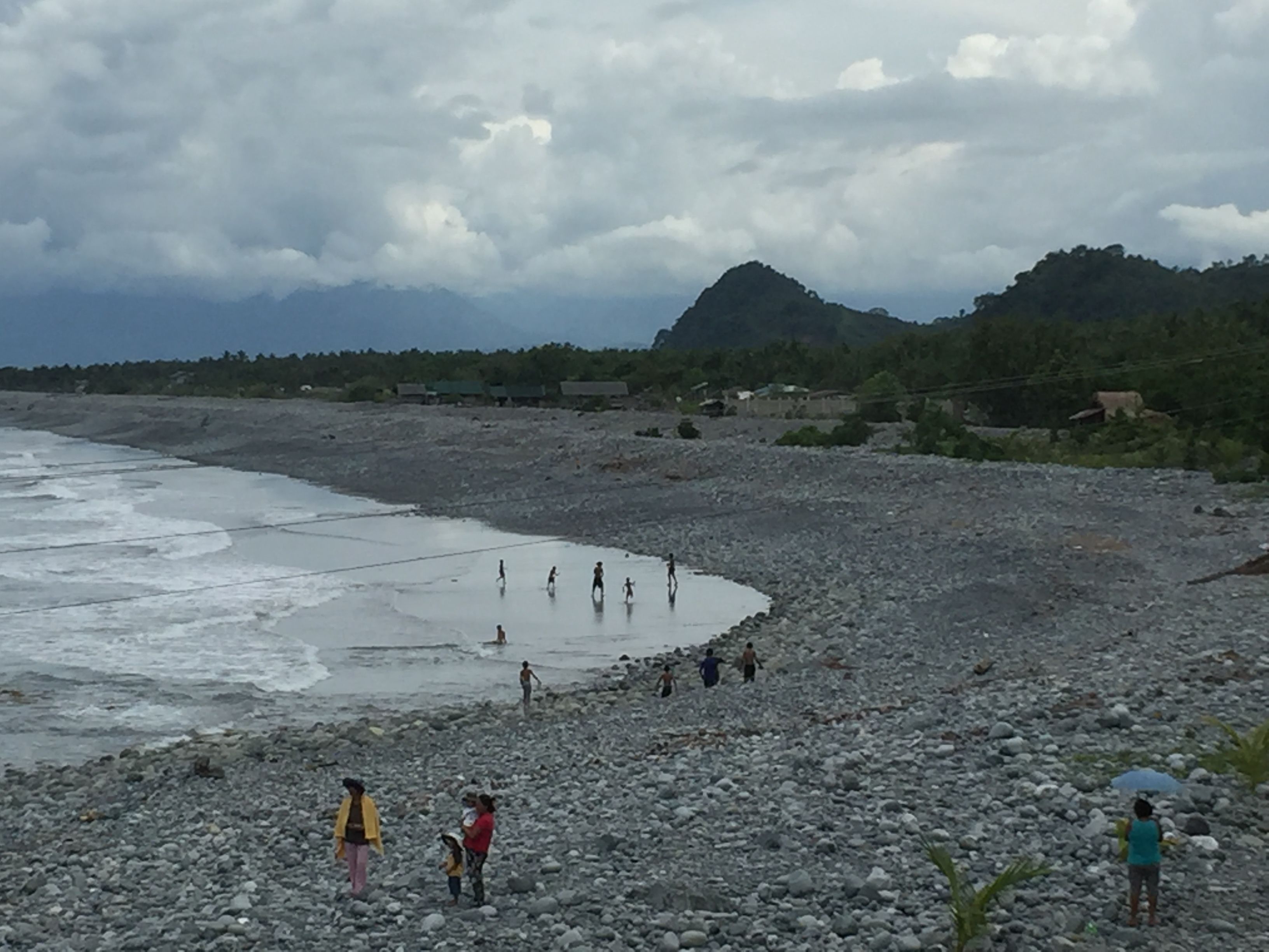 A rocky beach with people walking and standing, a mountain in the background, and a cloudy sky.