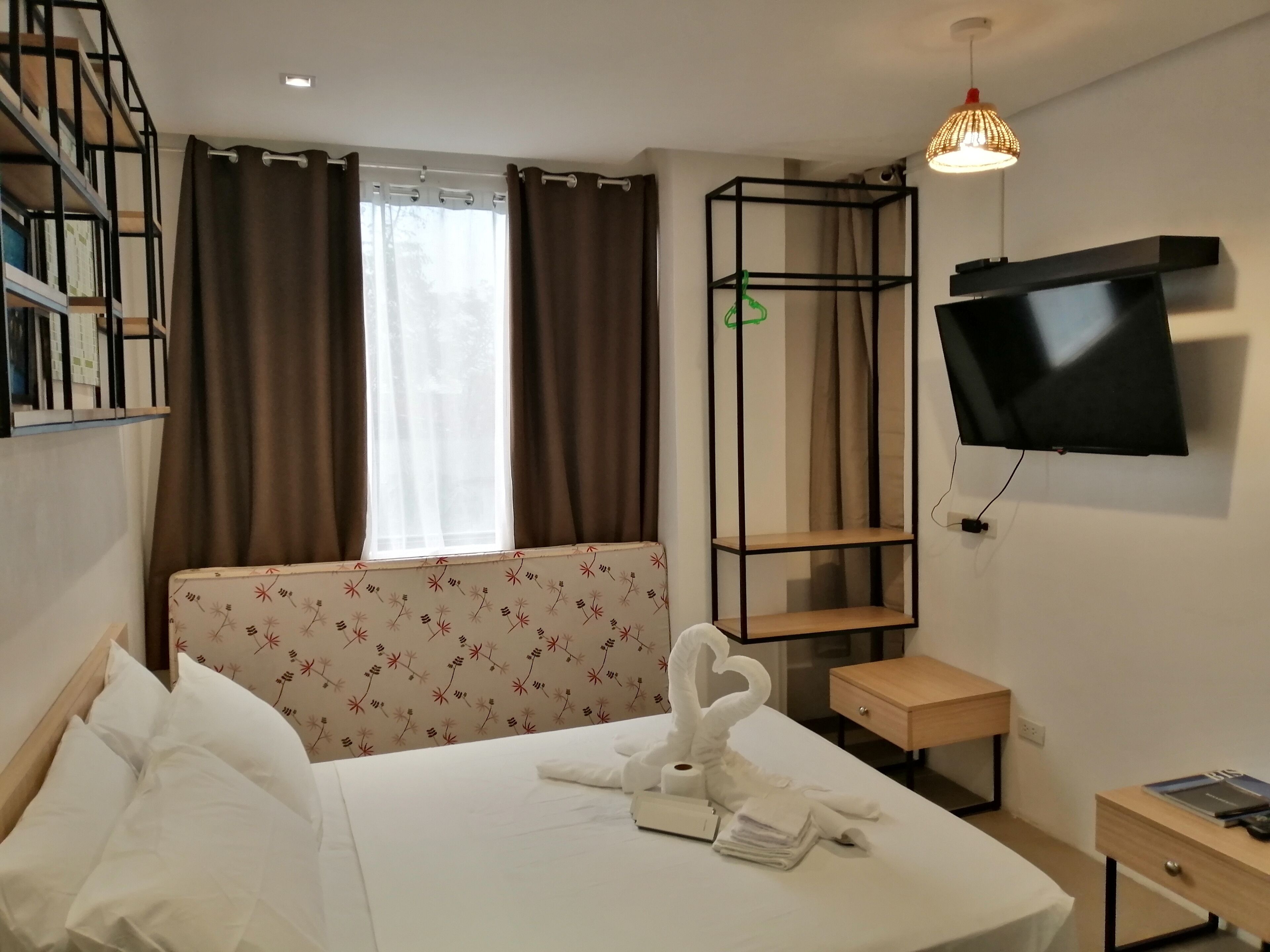 A hotel room with a bed, a TV mounted on the wall, a small wooden nightstand, a desk, and a window with curtains.