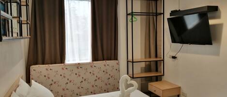 A hotel room with a bed, a TV mounted on the wall, a small wooden nightstand, a desk, and a window with curtains.