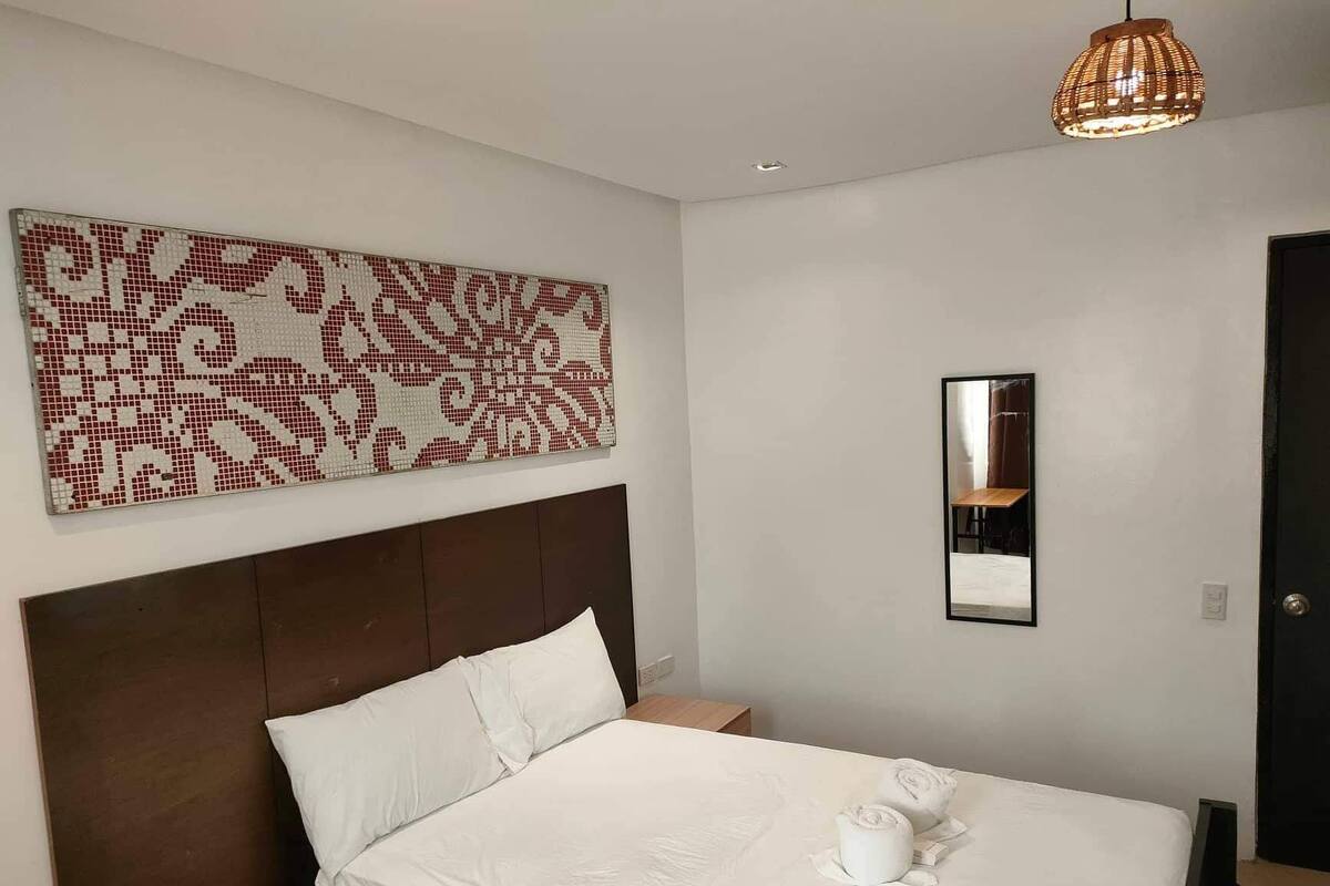 A hotel room with a large bed, a headboard, a wall-mounted artwork, a bedside table with a bottle and a tissue, and a hanging light fixture.