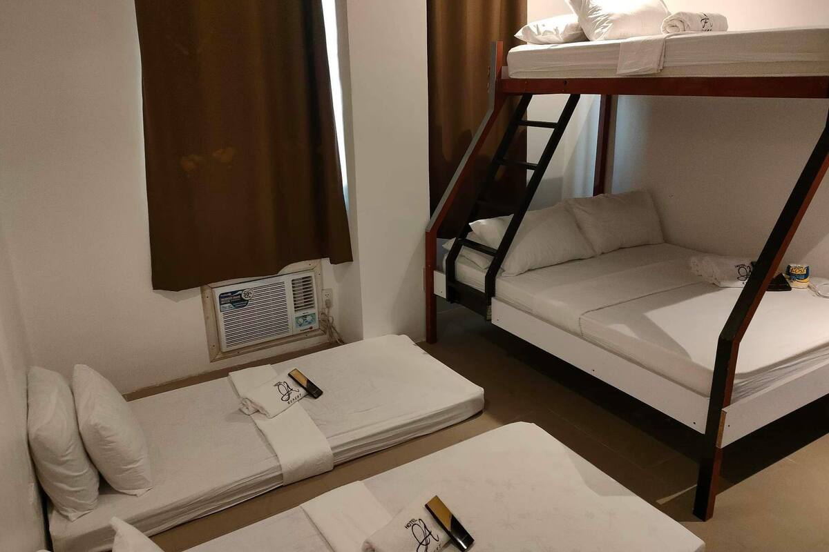 A compact hotel room with a bunk bed, a single bed, and a small table with a bottle and glasses.