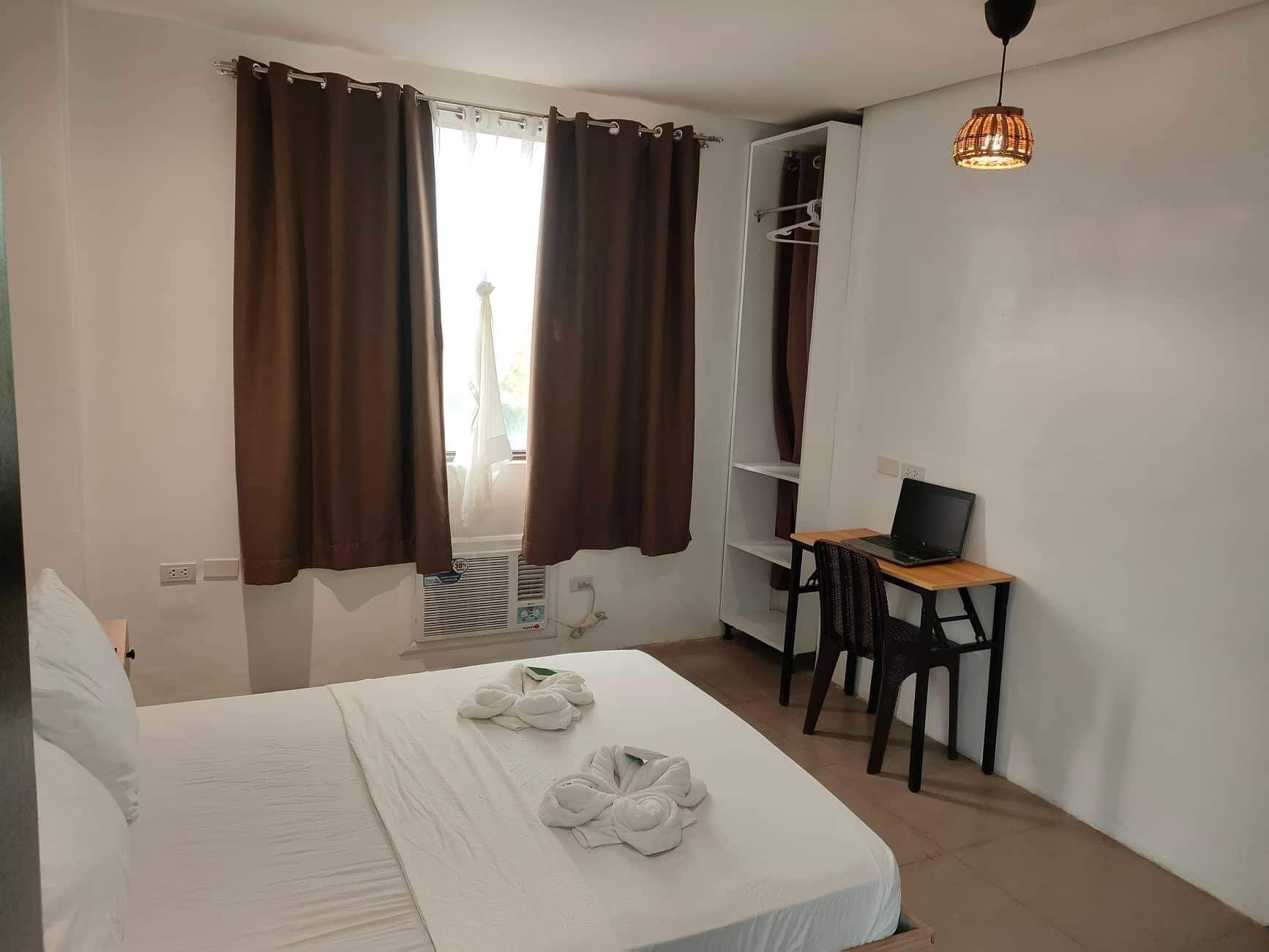 Hotellihuone, jossa on sänky, työpöytä kannettavalla tietokoneella, vaatekaappi ja riippuvalaisin.