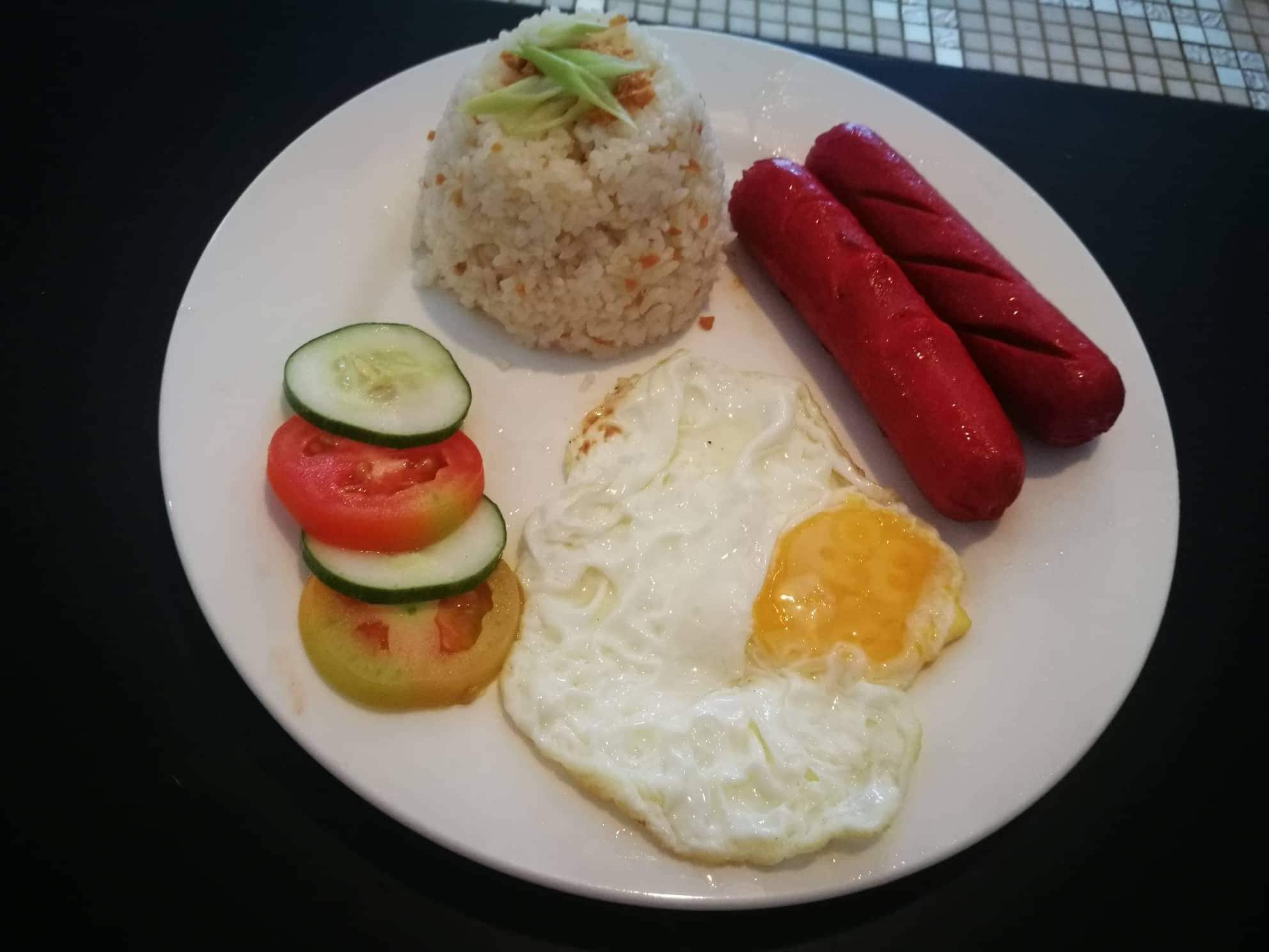 Un plato con arroz, dos salchichas, un huevo frito y verduras en rodajas.