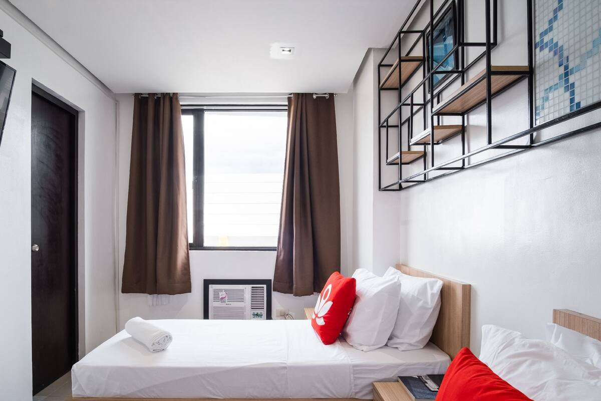 A hotel room with a bed, a TV mounted on the wall, a window with curtains, and a built-in shelf with a mosaic backsplash.