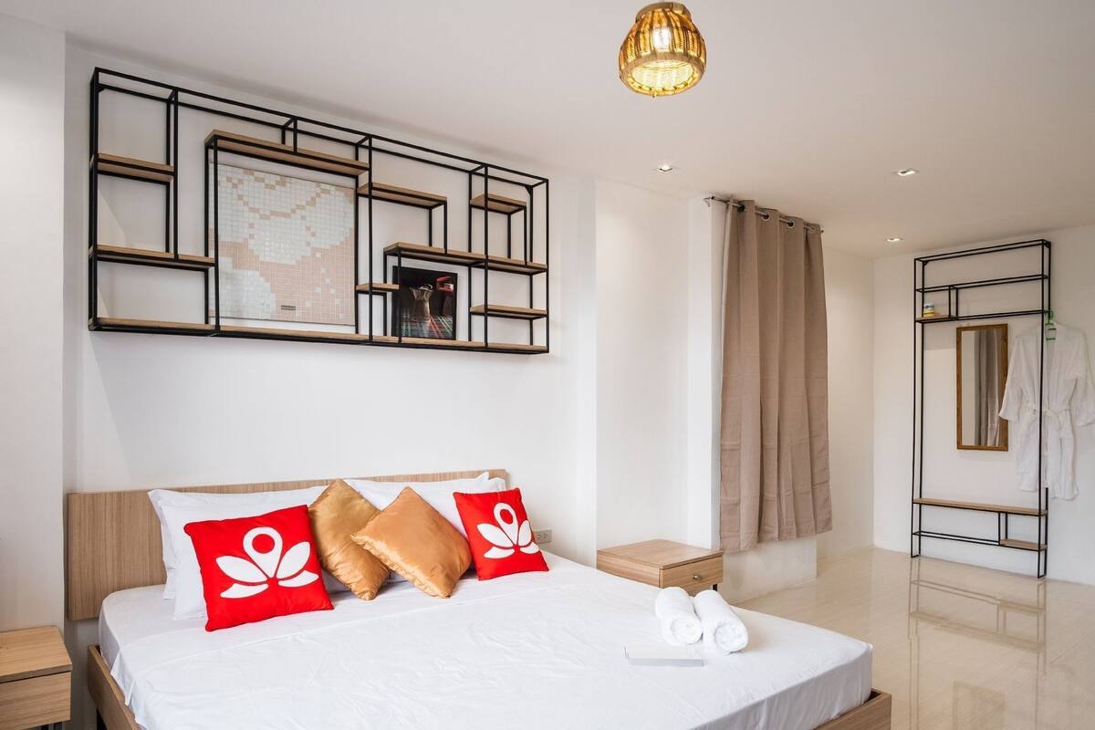 A hotel room with a bed, bedside table, hanging robe, and a wall-mounted shelf with a decorative tile.
