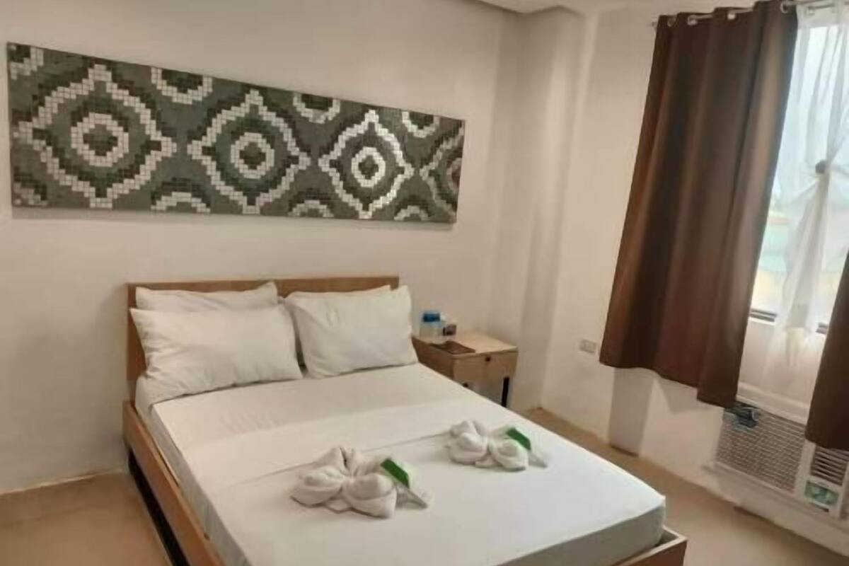 A hotel room with a bed, pillows, a nightstand, a wall-mounted air conditioner, and a window with curtains.