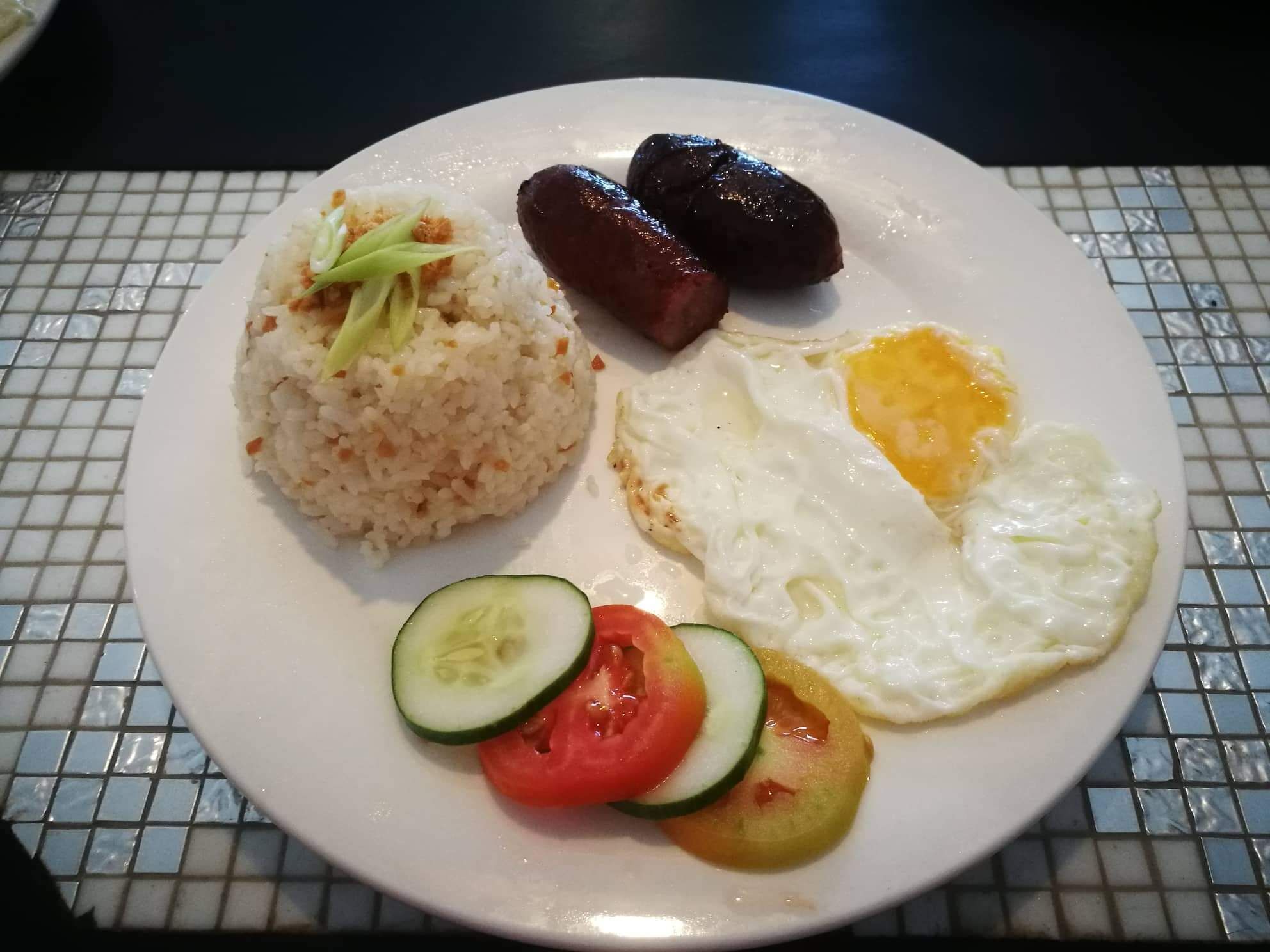 A plate with rice, sausages, fried eggs, and sliced vegetables.