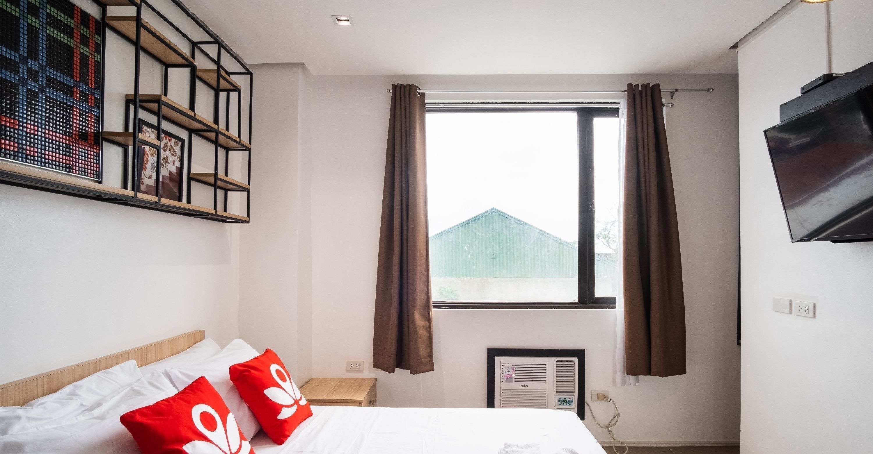 A hotel room with a bed, a TV, a window with curtains, a wall-mounted shelf with pictures, and a small table.