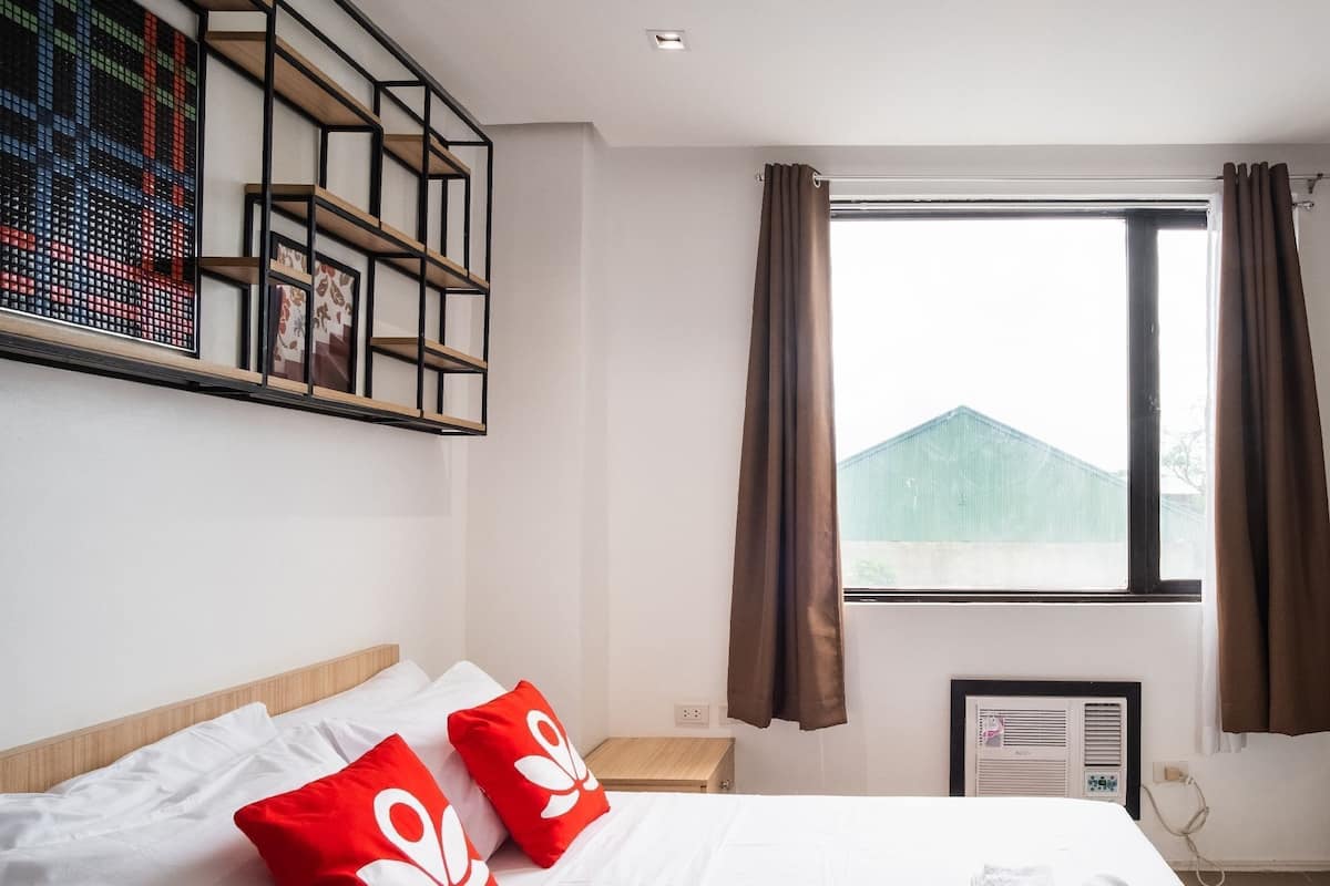 Habitación de hotel con cama, televisor, ventana con cortinas, estante en la pared con fotografías y una pequeña mesa.