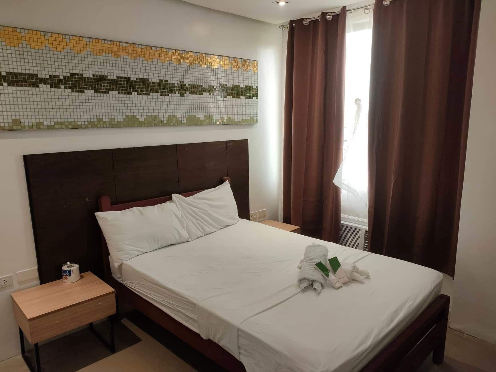 Una habitación de hotel con una cama, una mesita de noche, una pared con un mosaico y una ventana con cortinas.