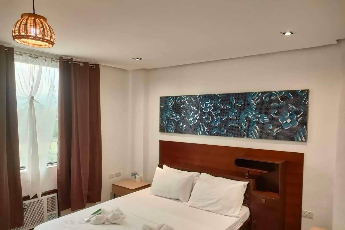 A hotel room with a large bed, a wooden headboard, a wall-mounted abstract art piece, a bedside table, and a window with curtains.