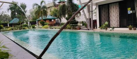 A swimming pool with clear water, surrounded by lush greenery and a building with a balcony.
