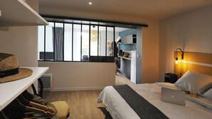 A hotel room with a bed, a desk, a laptop, a hat on the desk, and a view of the kitchen and balcony. - CHARMING APARTMENT WITH TERRACE IN A QUIET RESIDENCE NEAR JONZAC CENTER (Jonzac)