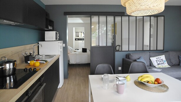 A modern kitchen with a microwave, stove, and utensils. There is a dining area with a table, chairs, and a fruit bowl. A gray sofa and a decorative pillow are visible in the background. - CHARMING APARTMENT WITH TERRACE IN A QUIET RESIDENCE NEAR JONZAC CENTER (Jonzac)
