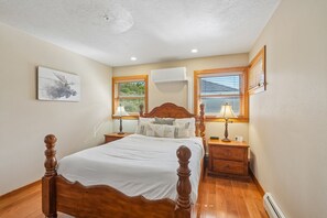 Une chambre à coucher avec un lit en bois, deux tables de chevet avec des lampes, un climatiseur fixé au mur et une fenêtre avec des stores.