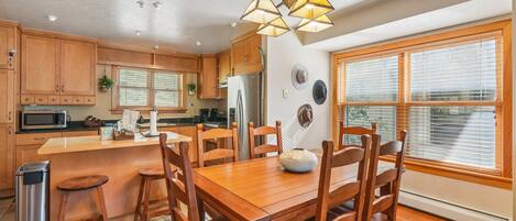 A kitchen with wooden cabinets, a central island, a dining table with chairs, a trash bin, and a large window with blinds.