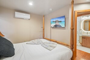 Une chambre d’hôtel avec un lit, un tableau représentant une plage tropicale, une tête de lit en bois et une salle de bain avec un miroir et un lavabo.