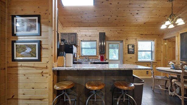A wooden cabin interior with a kitchen area featuring a large island with stools, a dining table, and framed pictures on the walls. - Firefly at Lazy River Cabins on Mt Fork River (Broken Bow)