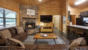 A cozy cabin interior with a stone fireplace, wooden walls, a leather sofa, a wooden coffee table, and a kitchen area with bar stools. - Firefly at Lazy River Cabins on Mt Fork River (Broken Bow)