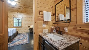 A wooden cabin interior with a bed, a bathroom with a sink and mirror, and a window with blinds. - Firefly at Lazy River Cabins on Mt Fork River (Broken Bow)