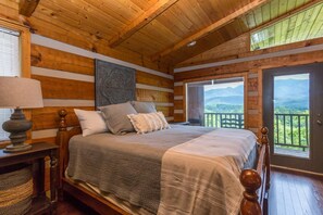 A wooden cabin bedroom with a bed, a nightstand, a lamp, and a view of mountains through a window. - Amazing Mt. LeConte Views/Gatlinburg Log Cabin (Gatlinburg)