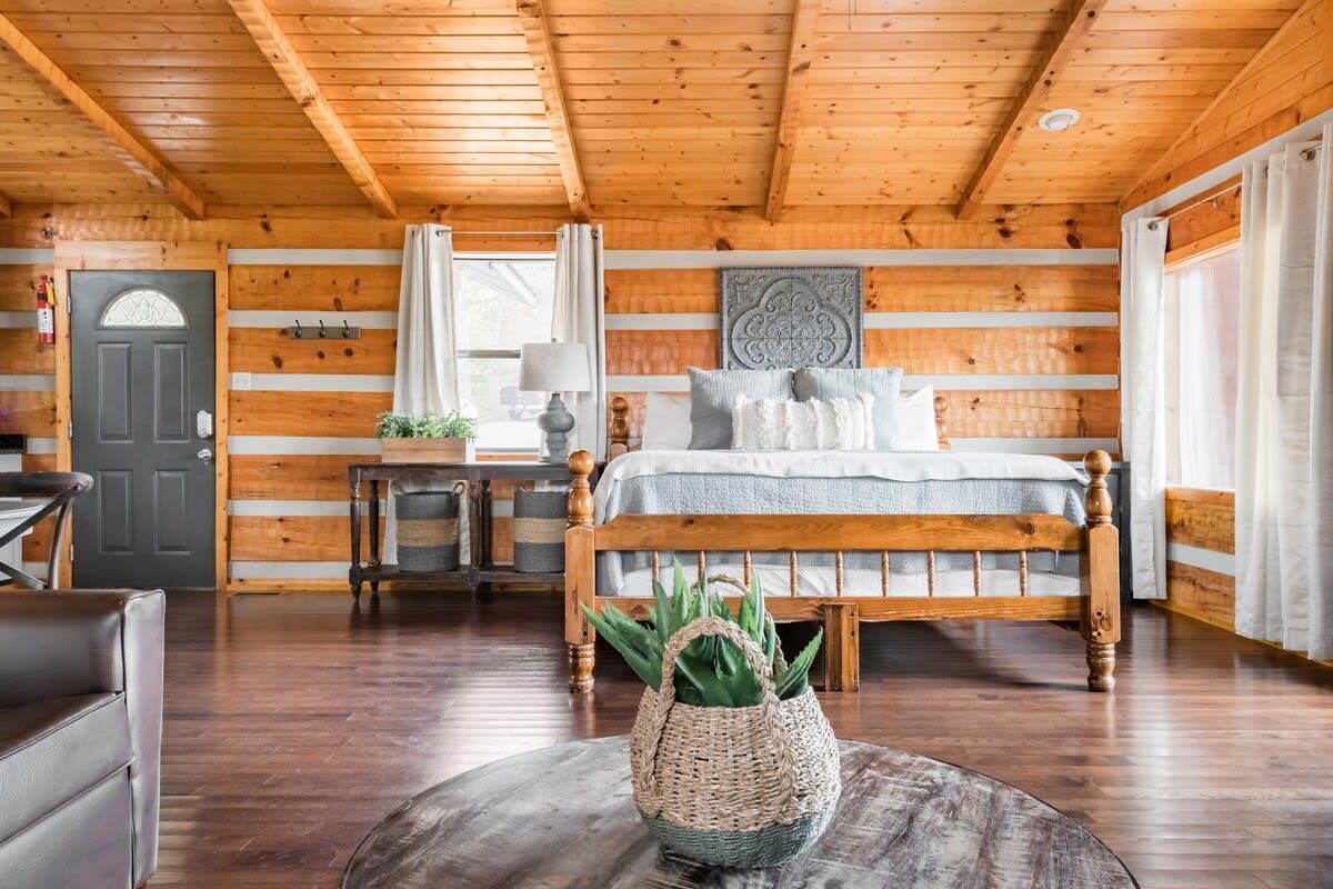 Un dormitorio tipo cabaña de madera con una cama de madera, una mesa redonda de centro con una cesta tejida y una puerta con ventanal.