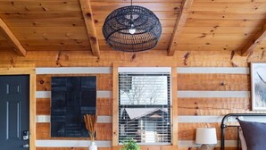 A wooden cabin interior with a round table, two chairs, a hanging light fixture, a window with blinds, and a bed with a headboard.