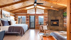 A log cabin interior with a bed, seating area, fireplace, and a ceiling fan.