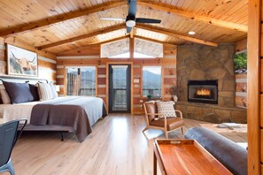 A log cabin interior with a bed, seating area, fireplace, and a ceiling fan.