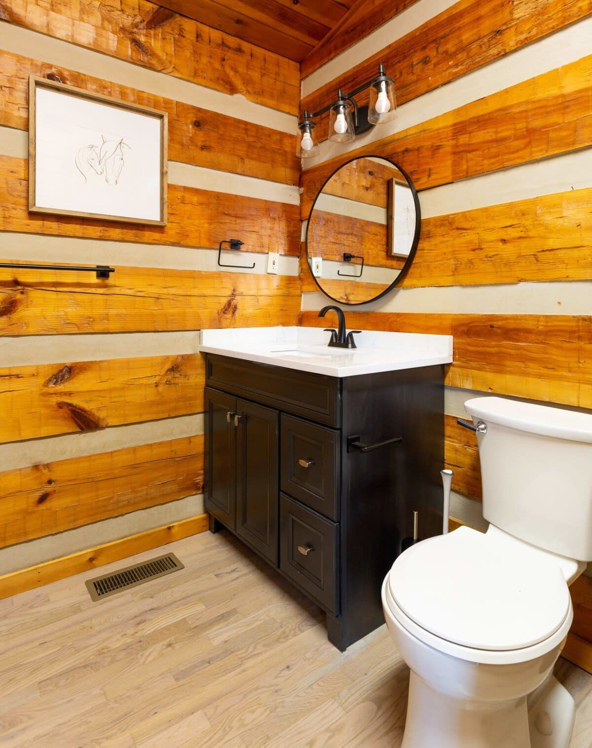 Un baño con paneles de madera, un lavamanos blanco, armarios oscuros, un espejo redondo y un inodoro blanco.