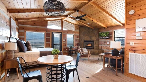 A cozy cabin interior with a wooden ceiling, a fireplace, a dining area with a round table and chairs, and a comfortable seating area with a sofa and chairs.