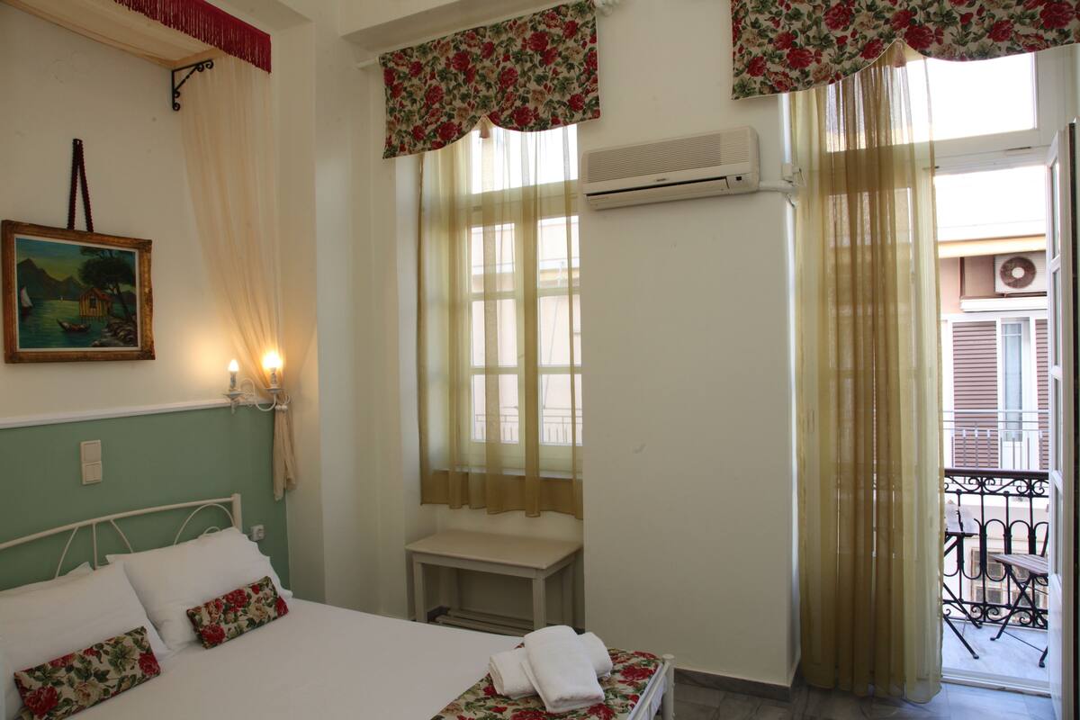 A bedroom with a bed, a bedside table, a window with curtains, an air conditioning unit, and a balcony with a railing.