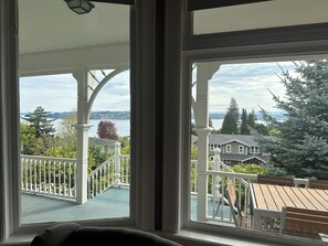 A room with a large window offering a view of a balcony, a wooden deck, and a distant body of water.