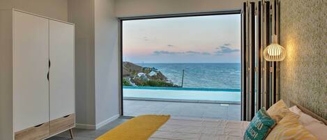 A bedroom with a large window overlooking the sea, a bed with yellow and patterned pillows, and a white wardrobe.