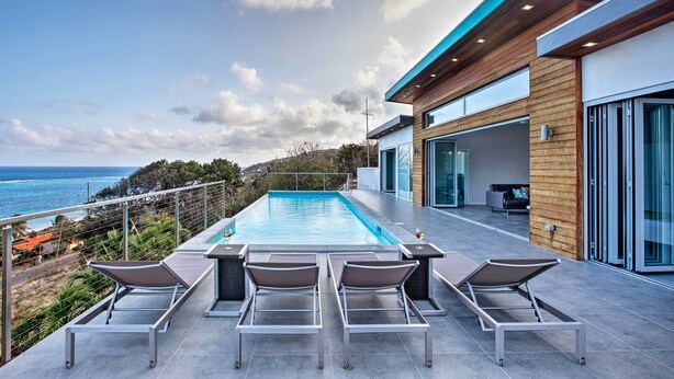 Un espace piscine extérieur moderne, doté de chaises longues, offrant une vue sur l’océan et comprenant un bâtiment contemporain aux grandes fenêtres.