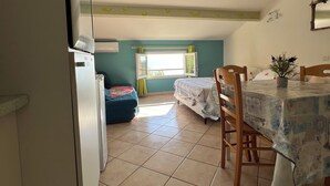 A room with a dining table, chairs, a bed, a window, and a refrigerator.