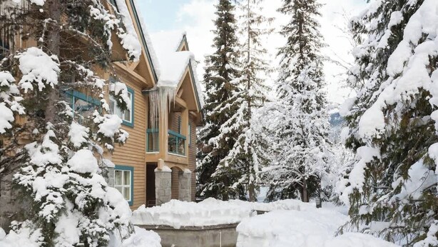 Eine Blockhütte mit verschneiter Fassade, umgeben von hoch aufragenden immergrünen Bäumen.