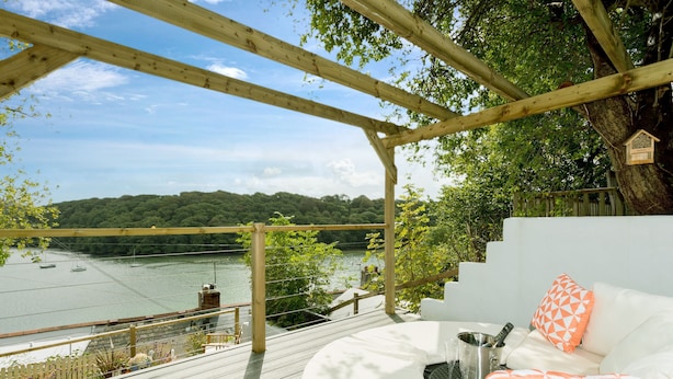 Une terrasse en bois avec un canapé blanc et des coussins orange, donnant sur une rivière et des collines boisées.